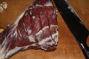 Para assar deliciosamente o cordeiro no forno, lave bem um pedaço de carne em água corrente e seque com uma toalha. Em seguida, faça vários cortes profundos na carne. Descasque o alho e corte-o em pedaços longitudinais. Coloque o alho nos cortes da carne.