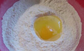 Sift the flour together with baking powder through a sieve and pour it into a deep bowl. Break a chicken egg in the center of the dry ingredient.