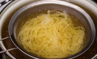 Commençons à assembler le plat. Placez les nouilles dans une passoire et faites-les chauffer dans un bouillon de bœuf, puis placez-les au fond d'un bol profond, garni de viande bouillie hachée, de racines d'oignon blanc, de rondelles de piment, de germes de soja et de feuilles d'herbes fraîches. 