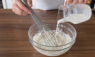 Now prepare the dough for pancakes. In a large container, sift the flour with soda and salt through a sieve. Then add granulated sugar and mix everything. Next, take a whisk and, without ceasing to stir, pour in milk and 250 ml of warm water.