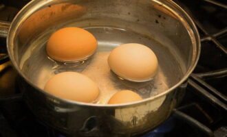 Place the eggs (take them out of the refrigerator in advance so they come to room temperature) into a saucepan and fill with warm water. Lightly add salt to the water. All these manipulations are necessary to ensure that the eggs do not crack during the cooking process. When the water boils, cook the eggs for about ten minutes until cooked. Then drain the boiling water and cool the eggs in cold water.