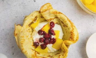 Take a small bowl and place the pancake so that the edges hang down. Place a couple of tablespoons of mousse, some peaches and boiled cherries in the cavity. We collect the pancake in a bag, leaving the ends on top. Using the same principle, fill the remaining pancakes, leaving one for the top of the cake.